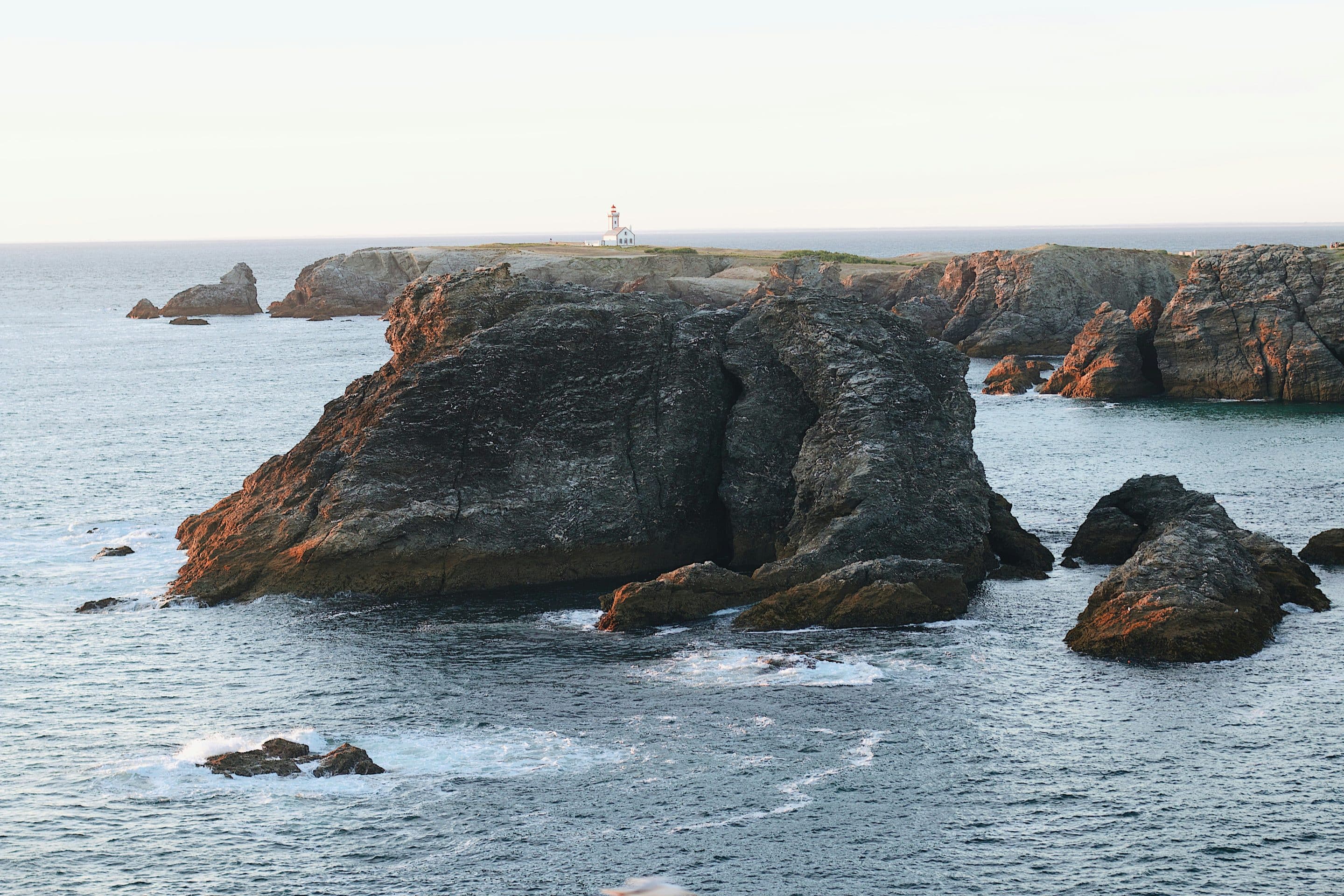 Paysage marin au lever de soleil à Belle-Ile-en-Mer avec des rochers et un phare à l'arrière-plan. 