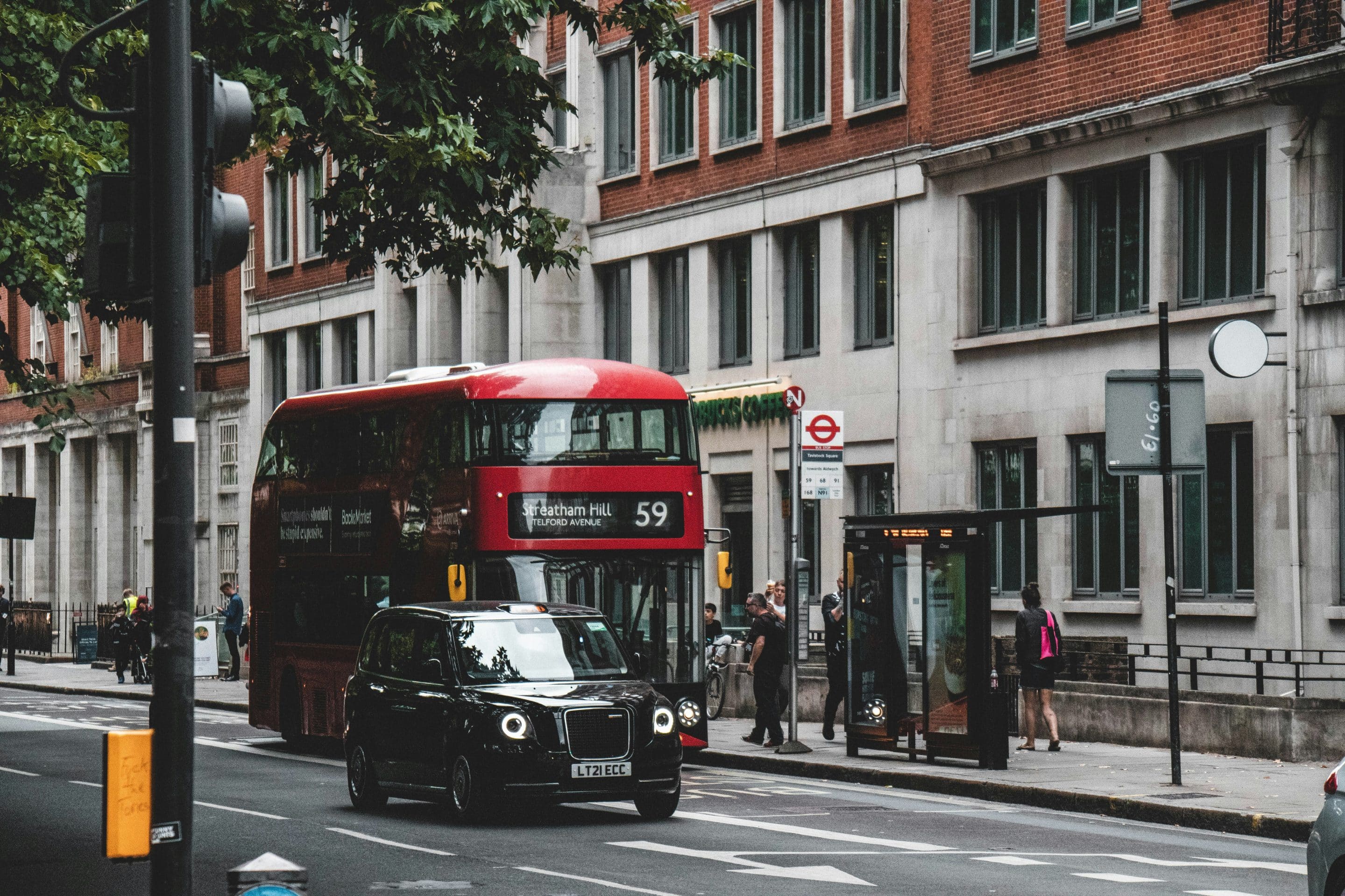 Rue londonienne avec un taxi et un bus à impérial. Des personnes sur le trottoir attendent pour monter dans le bus.