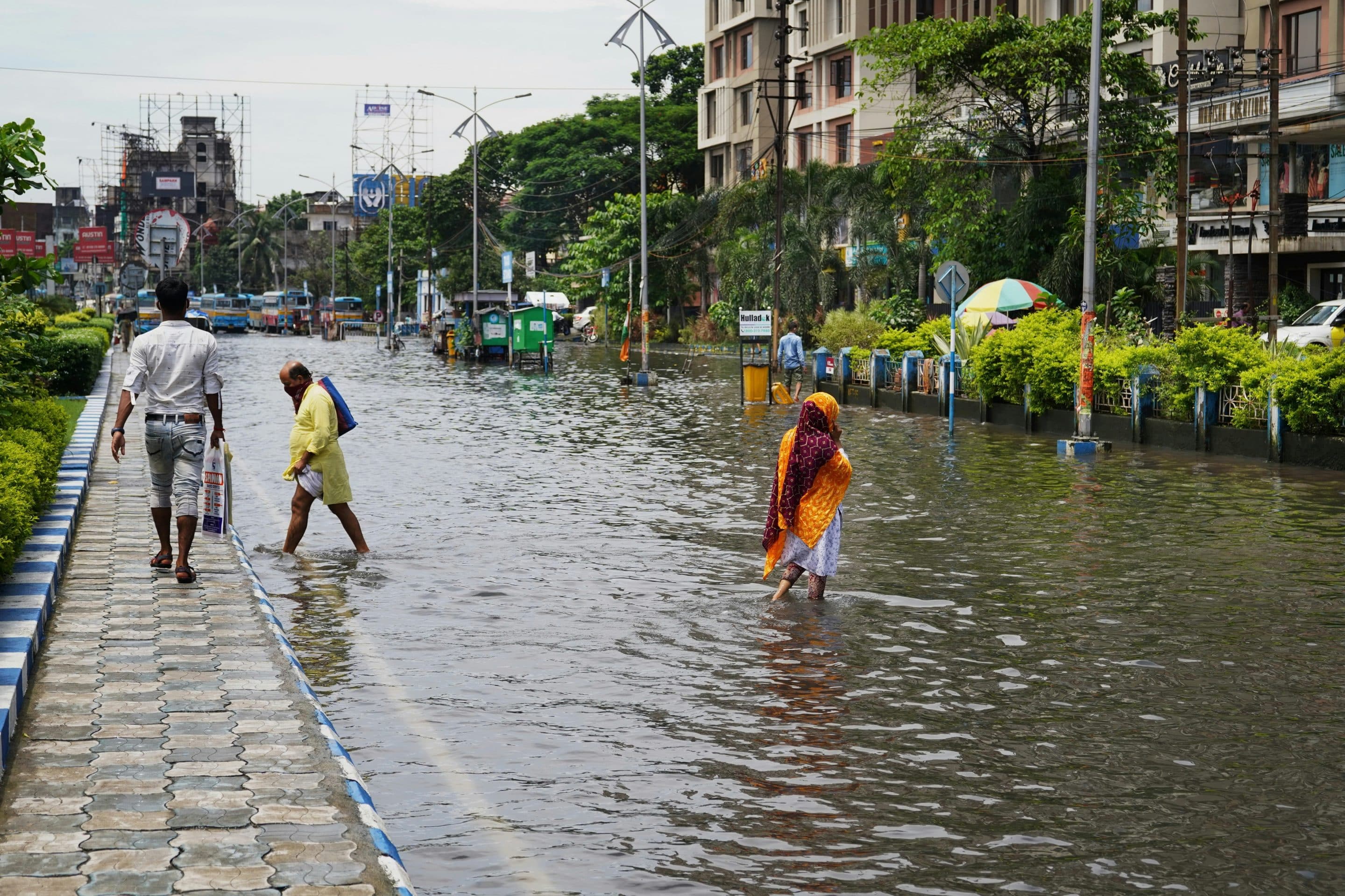Rue inondée avec bâtiments et espaces verts sur les côtés. Des personnes traversent les rues les pieds dans l'eau.